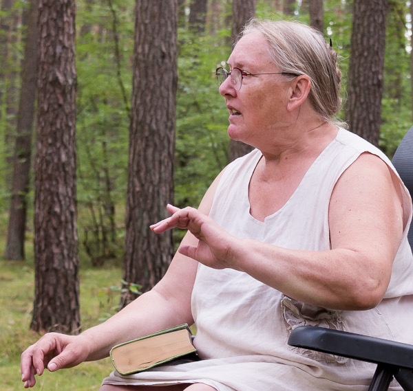 Karin sitzt auf einem Stuhl im Wald und erzählt. Hinter ihr sieht man Kiefernstämme und junge grüne Bäume. Sie gestikuliert mit ihrer linken Hand. Sie ist bekleidet mit einem ärmellosen Kleid.