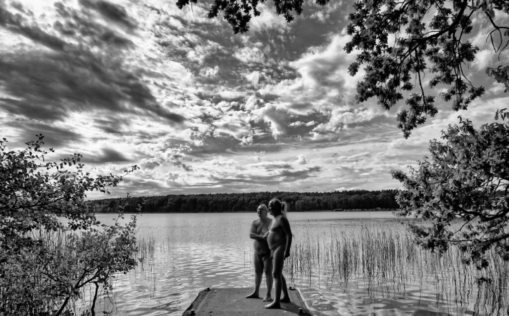 Wir stehen nackt auf einem Steg vor einem See, zwischen Laubbäumen und Schilf. In der Ferne sieht man das bewaldete andere Ufer und einen bewölkten Himmel.